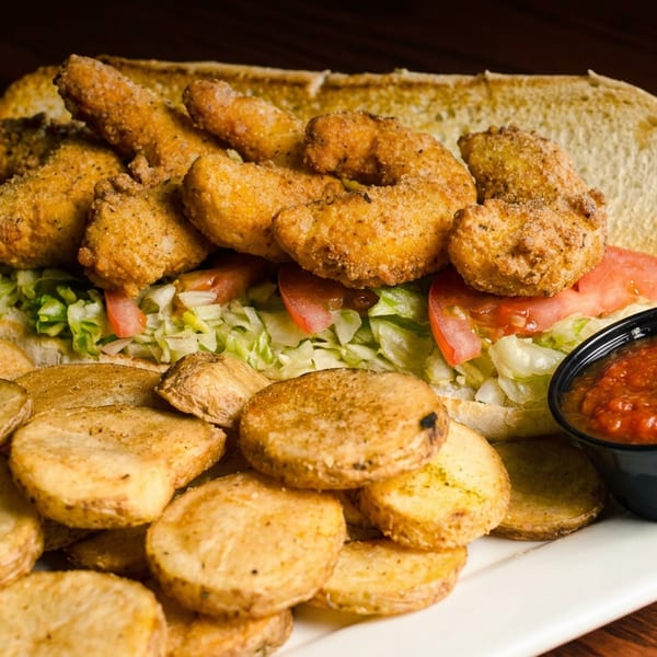 Fried seafood sandwich with lettuce and tomatoes, served alongside seasoned potato slices and red dipping sauce.