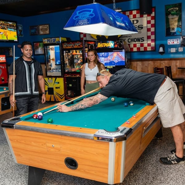 Three people play pool in a lively bar with arcade games, televisions, beer signs, and blue Bud Light billiard lights.