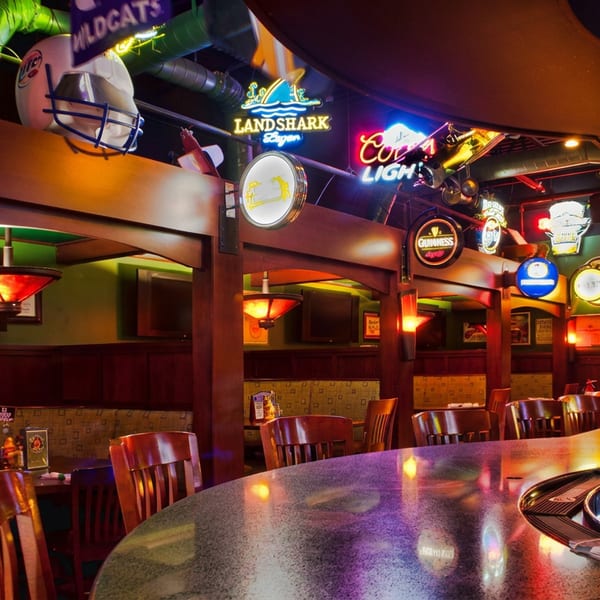 Colorful sports bar interior with a polished curved counter, beer taps, hanging glasses, booth seating, and neon beer signs.