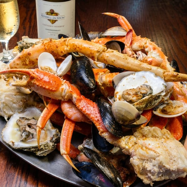 Seafood platter piled with crab legs, lobster, oysters, clams, mussels, and shrimp beside a glass of white wine.