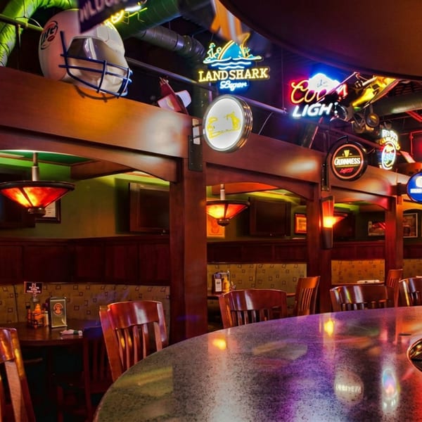 Dimly lit sports bar interior with wooden booths, empty tables, curved bar, colorful neon beer signs, and football memorabilia.