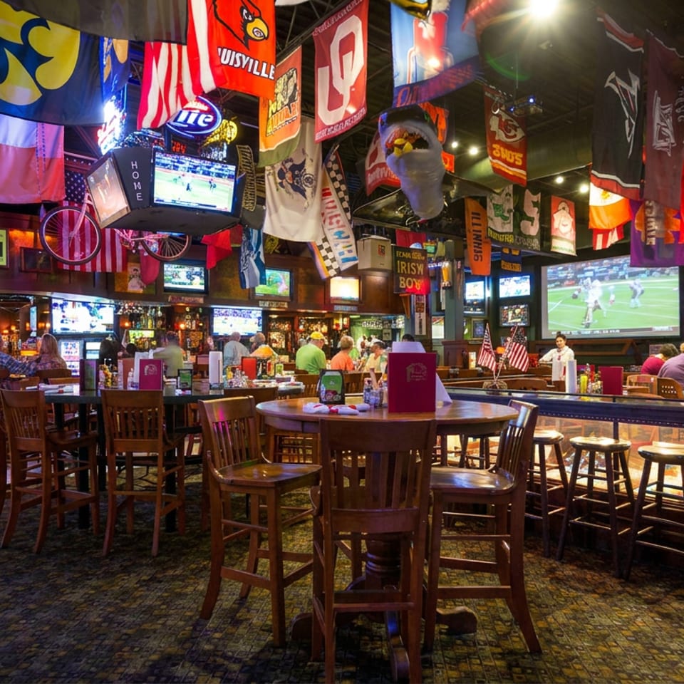 Lively sports bar filled with hanging team flags, numerous televisions showing games, wooden tables, and seated patrons.