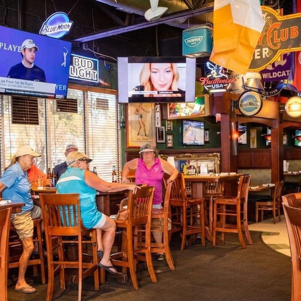 Sports bar interior with patrons seated at wooden tables, televisions showing sports, and colorful beer and team signs overhead.