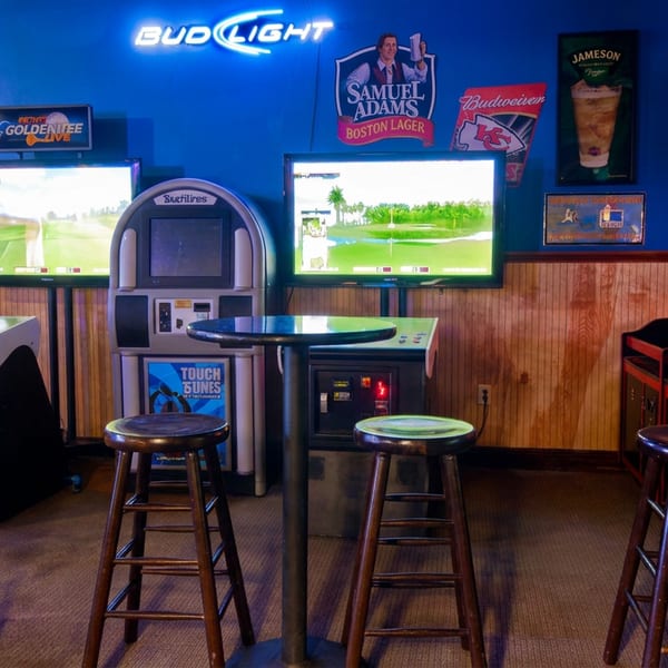 Sports bar game room with stools, golf simulator screens, arcade machines, beer signs, and a basketball game.