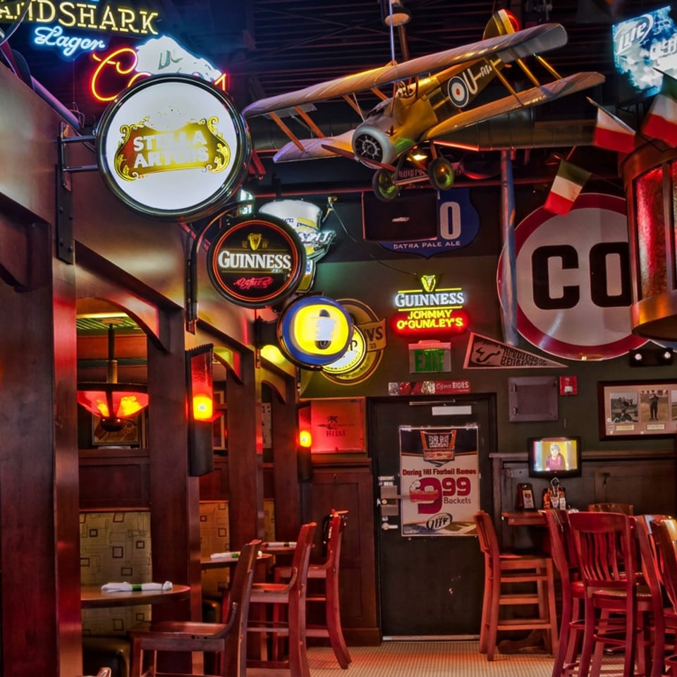 Dimly lit sports bar interior with red booths, wooden tables, beer signs, colorful neon lights, and a vintage airplane suspended overhead.