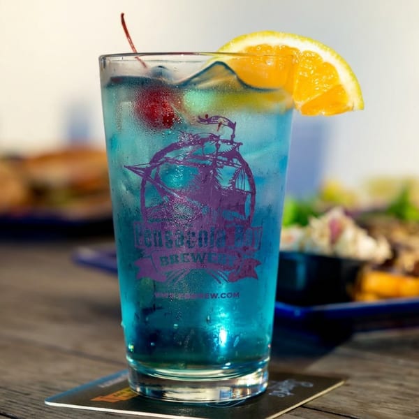 Blue cocktail garnished with an orange slice and cherry sits on a coaster, with plates of food blurred in the restaurant background.