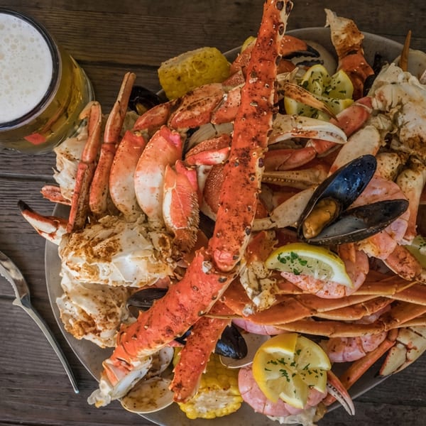 Overhead view of a seafood platter with crab legs, lobster, shrimp, mussels, corn, lemon, and a frothy beer on a wooden table.