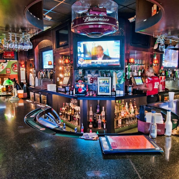Colorful sports bar interior with a polished counter, green lamps, hanging glasses, liquor bottles, televisions, and a bartender.