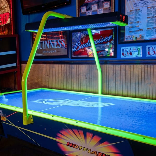 Neon-lit air hockey table in a sports bar, surrounded by beer advertisements, framed memorabilia, and arcade machines.
