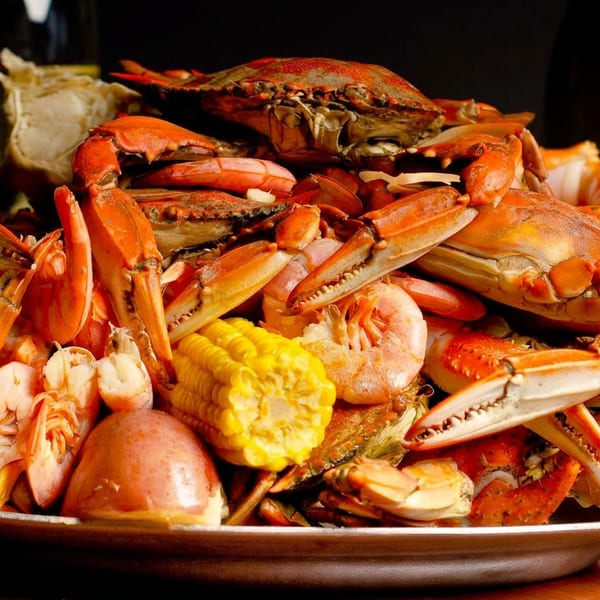Plate piled with steamed blue crabs, shrimp, lobster, corn, potatoes, and bread against a dark restaurant setting.