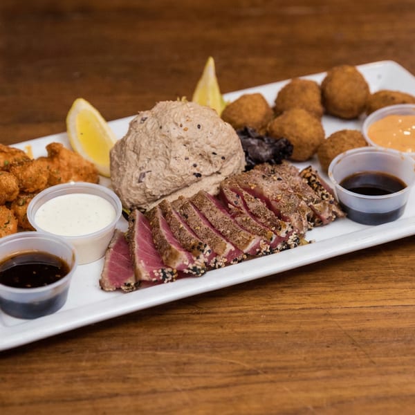 A white platter holds sliced seared tuna, fried bites, hummus, lemon wedges, and four dipping sauces on a wooden table.
