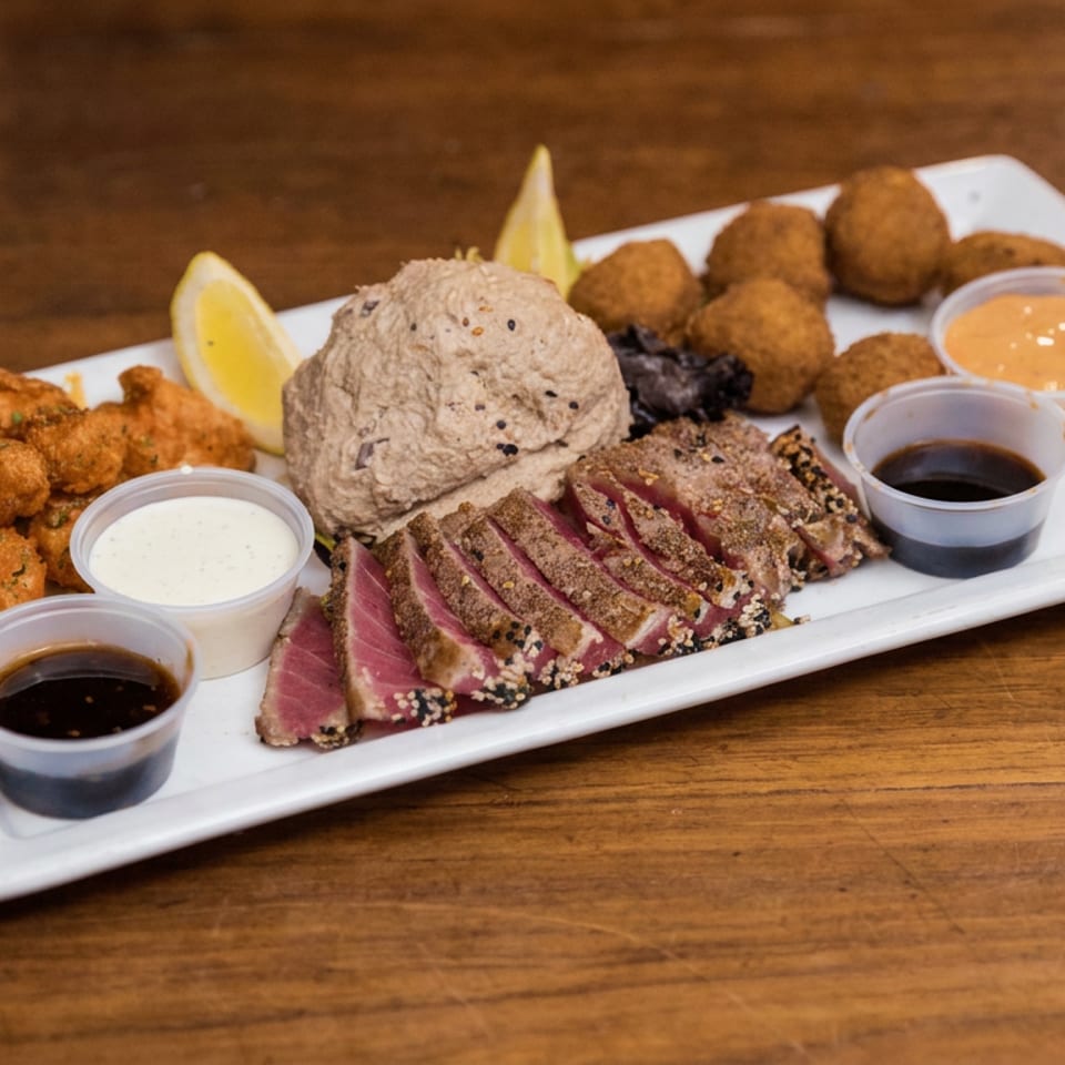 A white platter holds sliced seared tuna, fried bites, hummus, lemon wedges, and four dipping sauces on a wooden table.