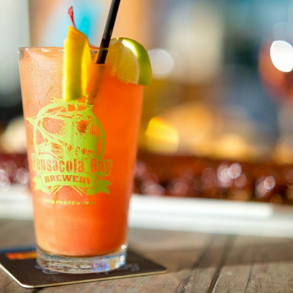 Bright orange cocktail garnished with citrus slices in a Gasparilla Bay Brewery glass, resting on a wooden restaurant table.