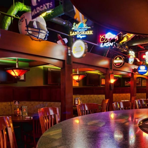 A lively bar interior with neon signs, sports memorabilia, and warm lighting over tables and a bar.