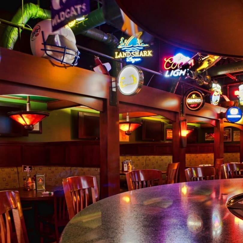 A lively bar interior with neon signs, sports memorabilia, and warm lighting over tables and a bar.