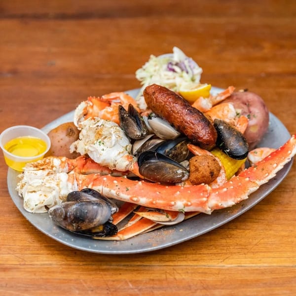 Seafood boil with crab legs, mussels, shrimp, sausage, potatoes, corn, and coleslaw on a metal platter.