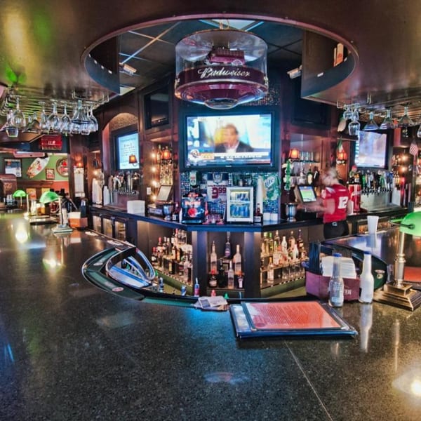 A wide shot of a bar with a bartender, bottles, glasses, and TVs showing sports.