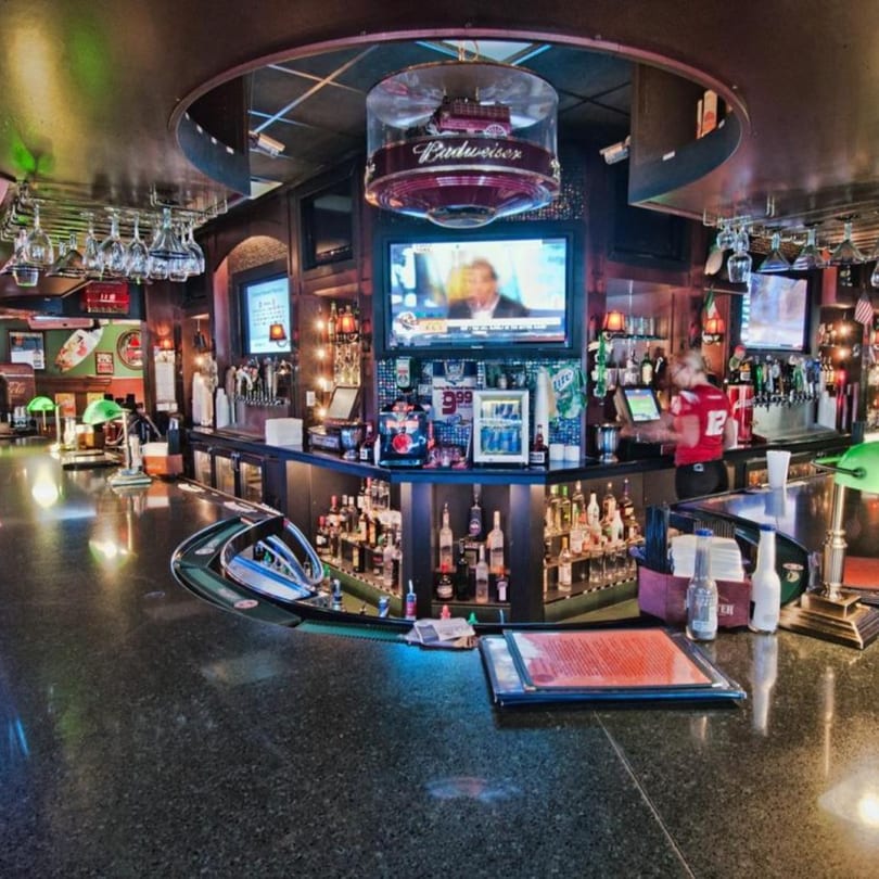 A wide shot of a bar with a bartender, bottles, glasses, and TVs showing sports.