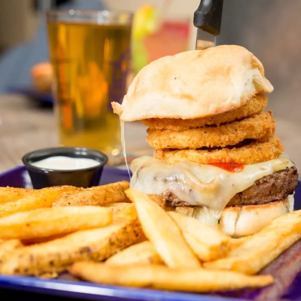 A towering burger with melted cheese, onion rings, and a knife, served with fries and a drink.
